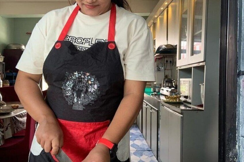 A person wearing a white shirt and a black apron with a red trim, standing in a kitchen with cabinets and a stove in the background. 4