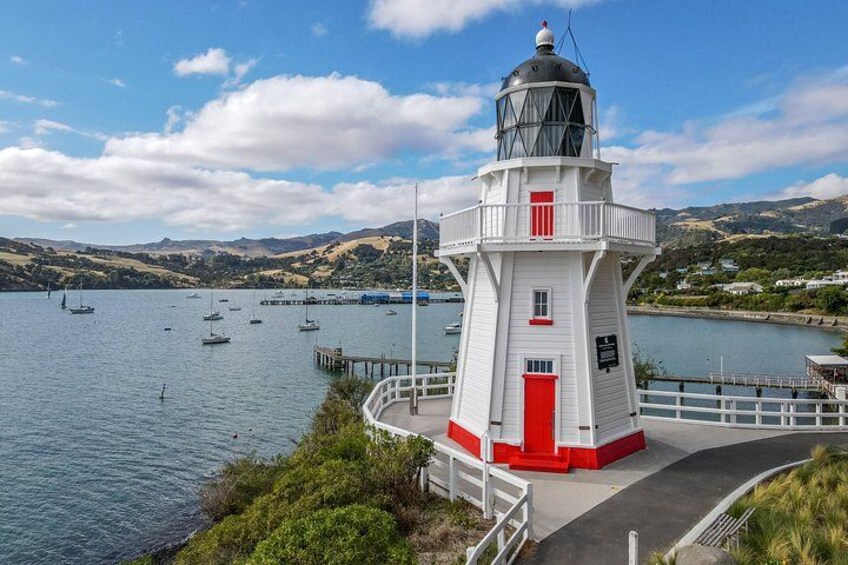 A lighthouse by a calm bay with boats, greenery, and hills in the background. 1