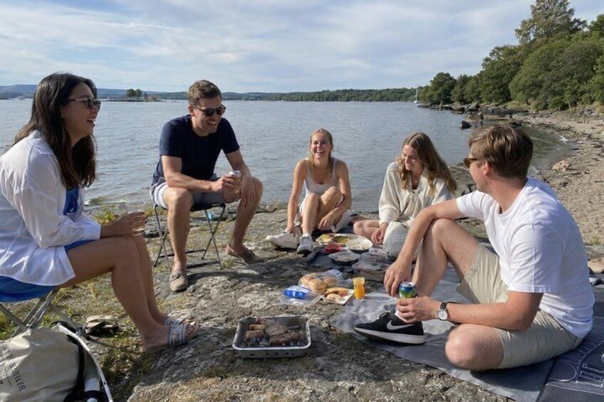 A group of people sitting on a rocky shore by a lake, with a portable grill and food items on the ground. 2