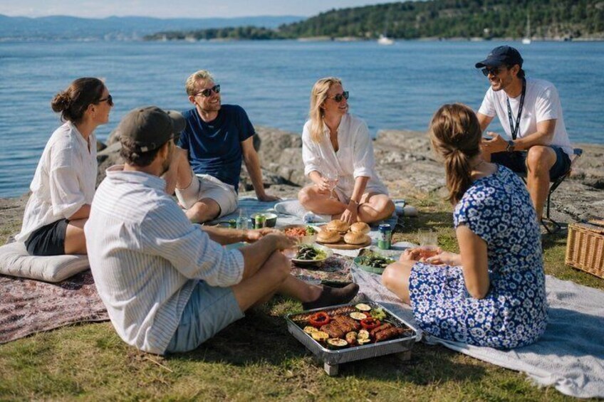 A group of people having a picnic by a lakeside with food and drinks on a blanket. 1