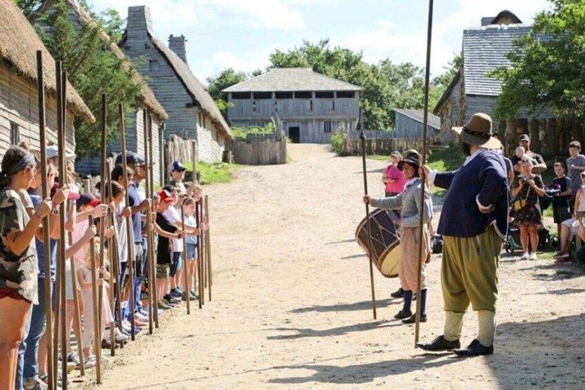 A historical reenactment with participants in period costumes, a dirt road, and thatched-roof buildings. 3