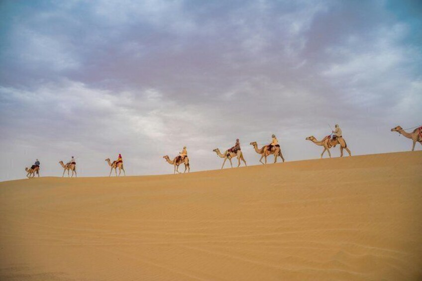 A group of people riding camels across a sandy dune landscape under a cloudy sky. 1