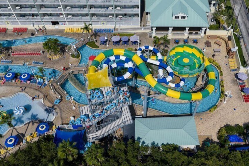 A water park with multiple slides and pools, surrounded by palm trees and a building with a green roof. 2