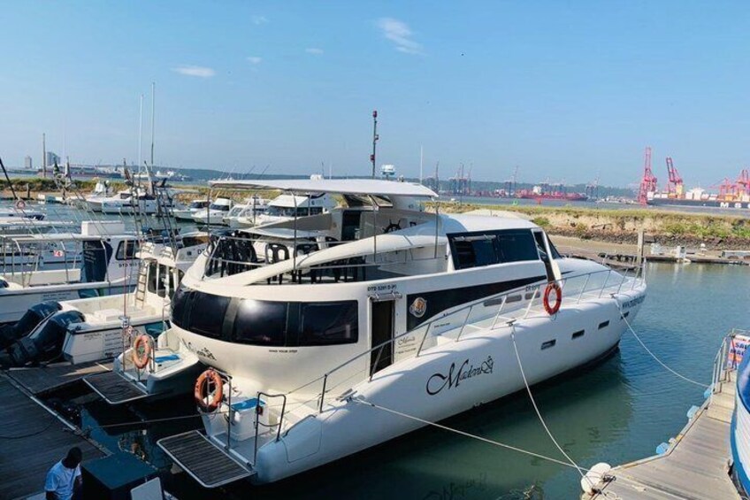 A white yacht docked at a marina with other boats, a clear sky, and industrial structures in the background. 2