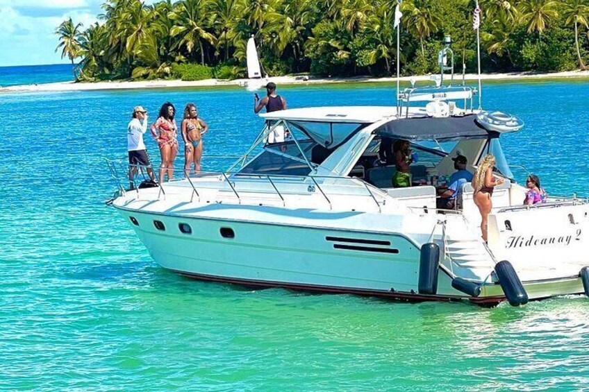 A white boat named 'Hidcana2' on turquoise water with people on deck and a beach with palm trees in the background. 1