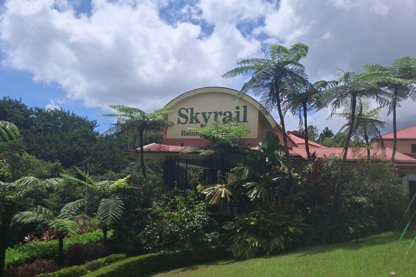 A sign for Skyrail Rainforest with lush greenery and a clear sky. 2