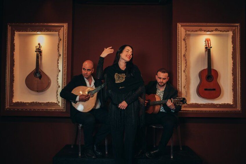 Three people seated on stools, two playing guitars, and one standing with a guitar, in a dimly lit room with framed guitars on the wall. 1