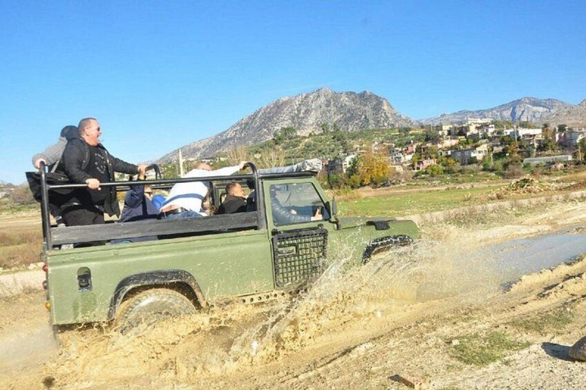 A green off-road vehicle with passengers on a dirt road, surrounded by a mountainous landscape and a village in the background. 5