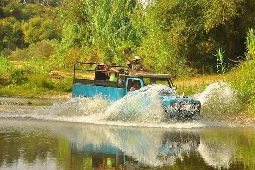 A blue off-road vehicle driving through water, splashing, surrounded by greenery. 1