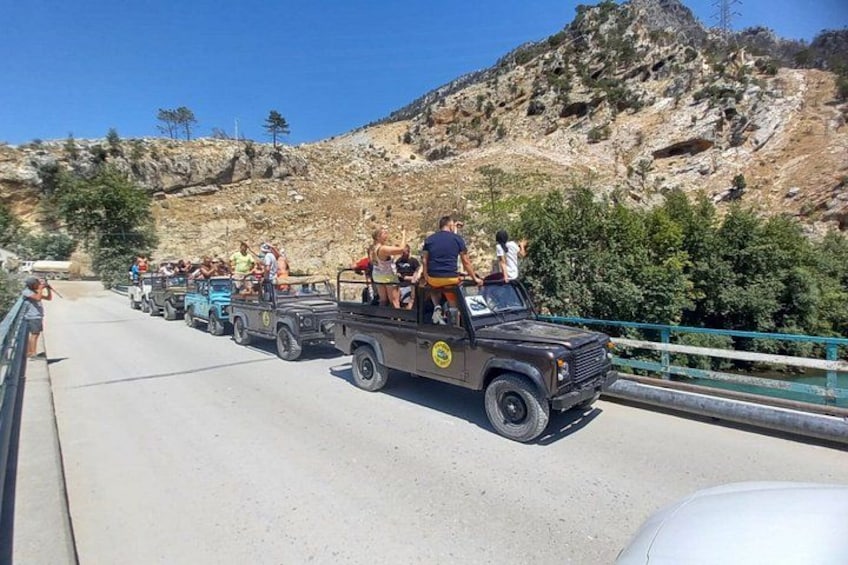A convoy of vehicles on a bridge with passengers, surrounded by rocky terrain and greenery. 4