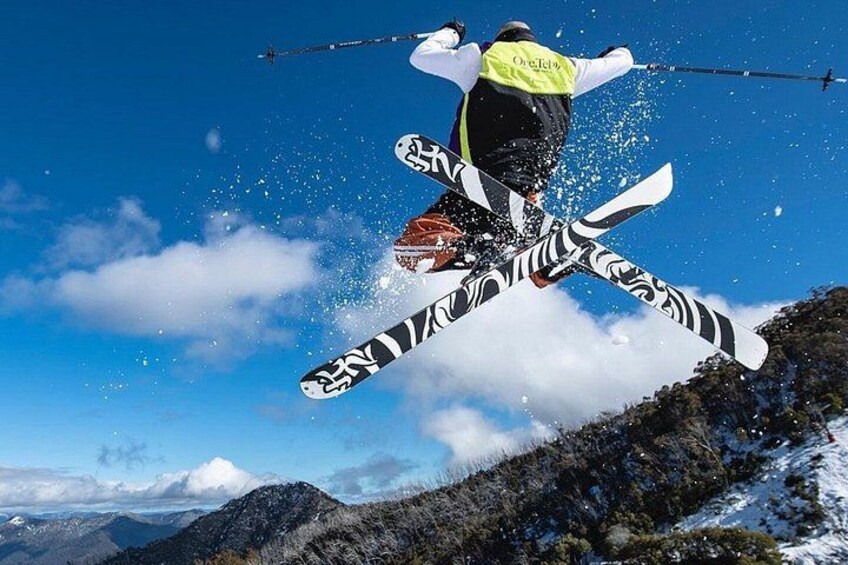 A skier in mid-air performing a jump with skis crossed, wearing a neon vest, against a mountainous backdrop. 2