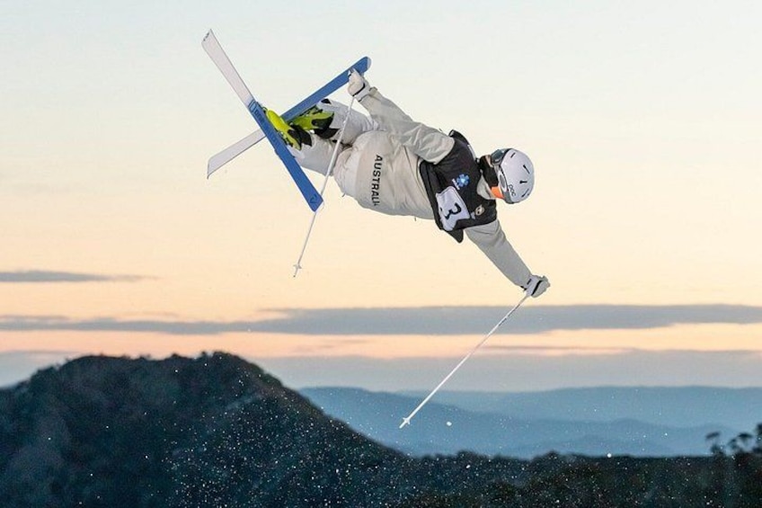 A skier in mid-air performing a jump with skis and poles, wearing a helmet and a numbered bib, against a mountainous backdrop. 1