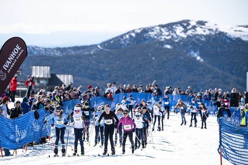 A cross-country skiing race with participants on a snowy trail, spectators behind barriers, and a mountainous backdrop. 5