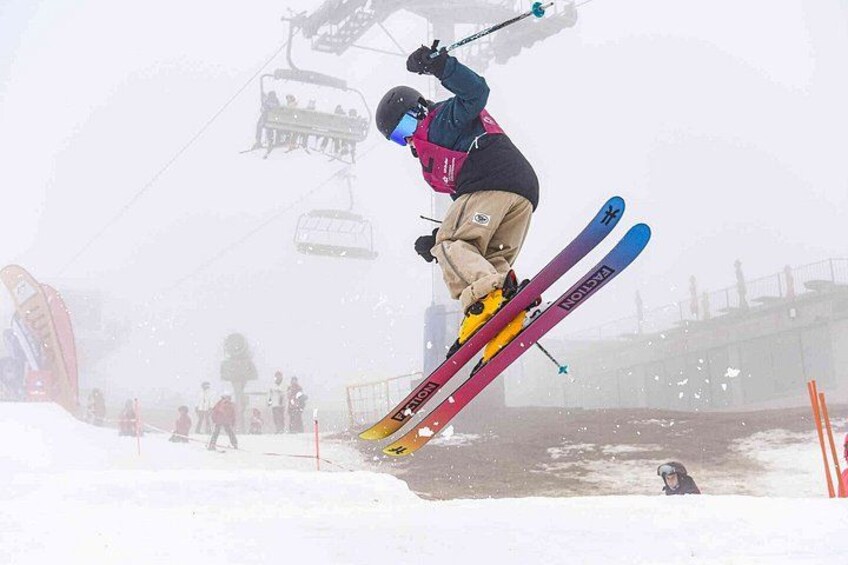 A skier in mid-air performing a jump, with a ski lift and spectators in the background. 4