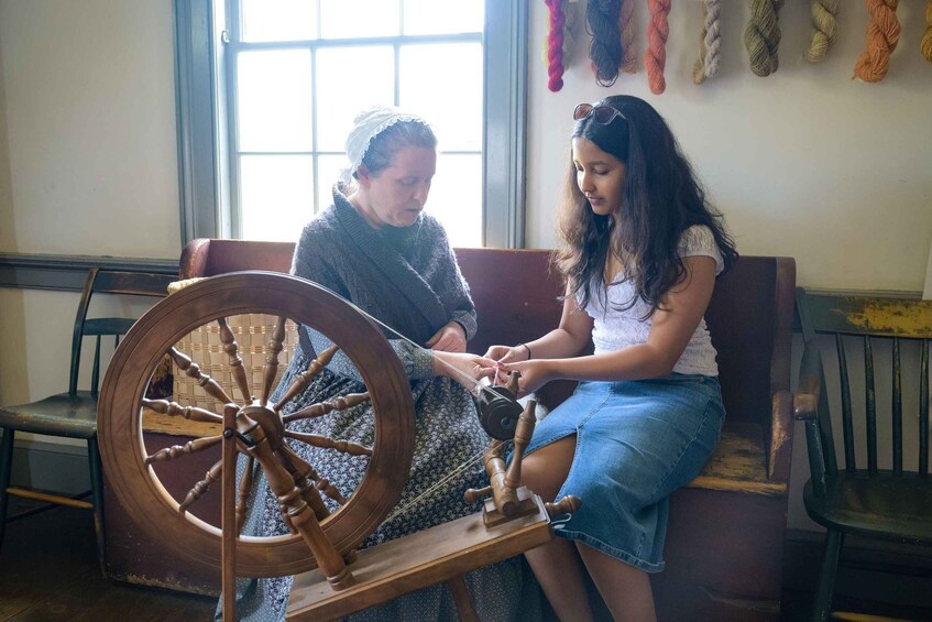 Two people sitting on a bench, one operating a spinning wheel. 4