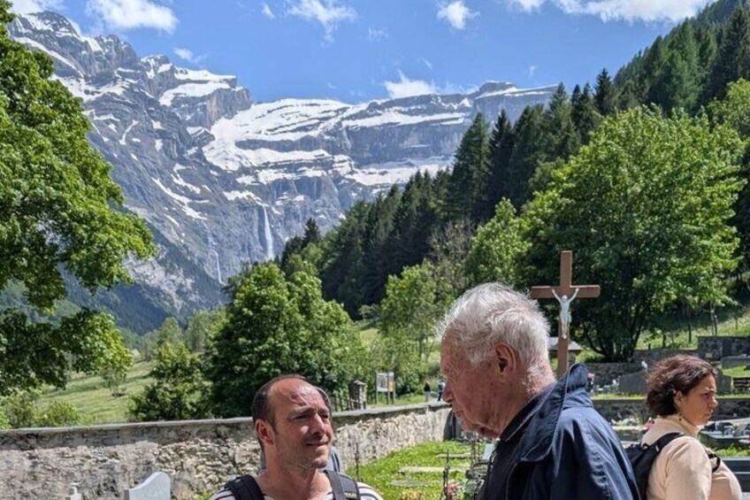A mountainous landscape with a cross, a person in a blue jacket, and a person in a beige jacket. 1