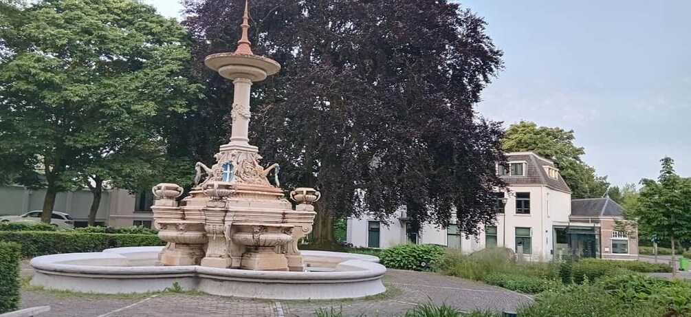 A classical fountain with a tall spire, surrounded by a circular stone basin, set in a park with lush greenery and a building in the background. 2