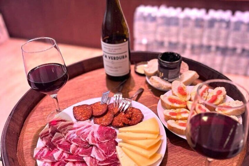 A wooden tray with a bottle of wine, a glass of red wine, a plate of cured meats, cheese, bread, and a small jar of condiment. 1