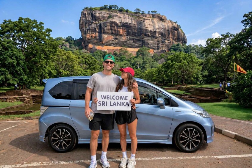 A couple standing in front of a car with a sign that reads 'WELCOME SRI LANKA' and a large rock formation in the background. 1