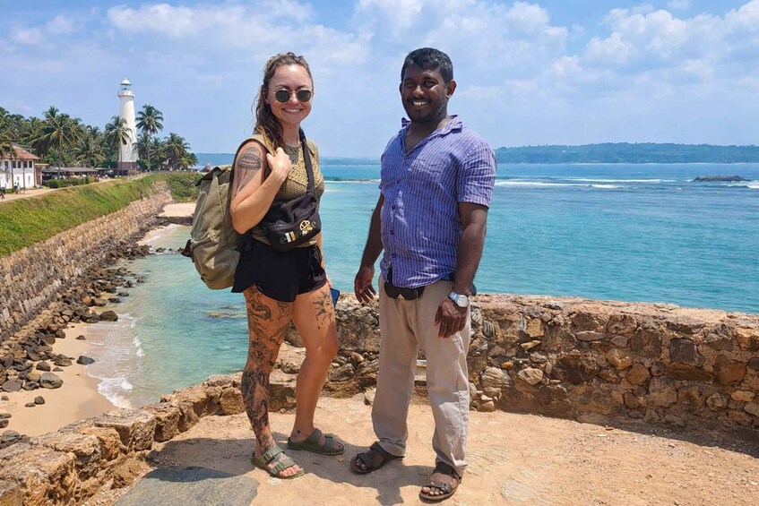 Two people standing by a coastal road with a lighthouse in the background. 4