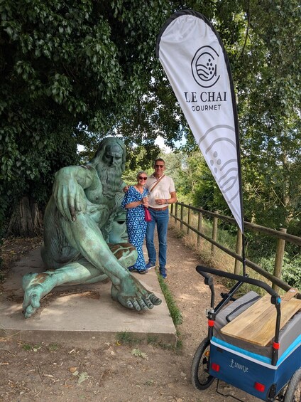 A bronze statue of a reclining figure, a white banner with 'LE CHAI GOURMET' text, and a blue and black bicycle cart. 3