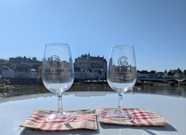 Two wine glasses with 'LE CHAI GOURMET' on a table by a river with buildings in the background. 1