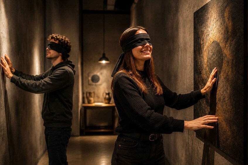 Two people in a dimly lit corridor, one blindfolded and the other reaching out to touch a textured wall. 1