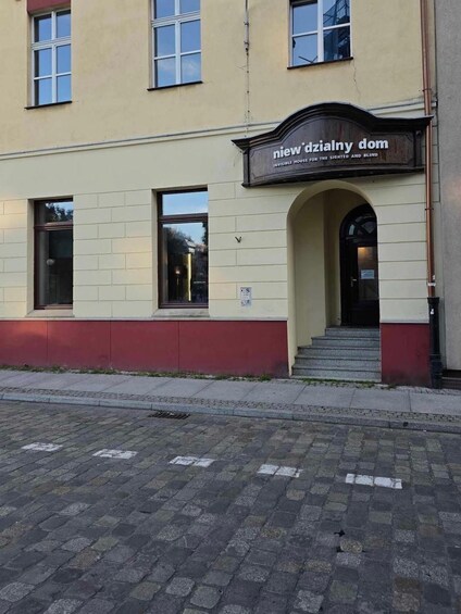 A building with a sign reading 'Nowe dzialny dom' and a red brick pavement in front. 2