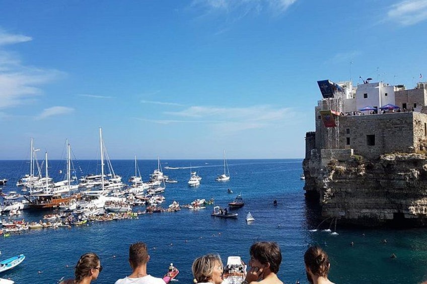 Un pueblo costero con un edificio histórico de piedra, un puerto deportivo con veleros y gente disfrutando del paseo marítimo. 4