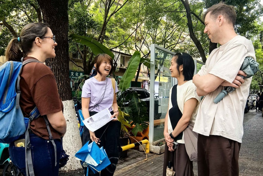 A group of people standing outdoors, some holding bags, with trees and a building in the background. 2