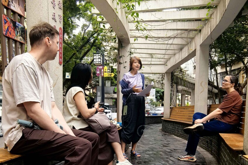 A group of people sitting and standing in a covered outdoor area with a brick floor, surrounded by greenery and a building with signage. 1