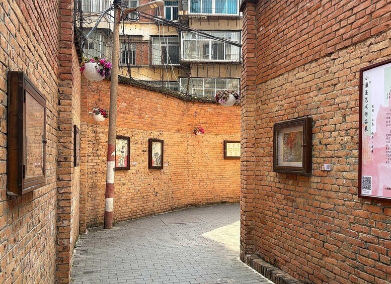 A narrow alley with brick walls, hanging flower baskets, and framed artwork. 5