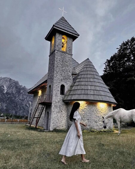 A stone church with a bell tower, a woman in a white dress, and a white horse in a grassy field. 5