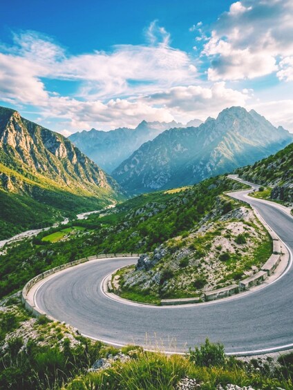 A winding mountain road surrounded by lush green hills and towering mountains under a clear blue sky. 2