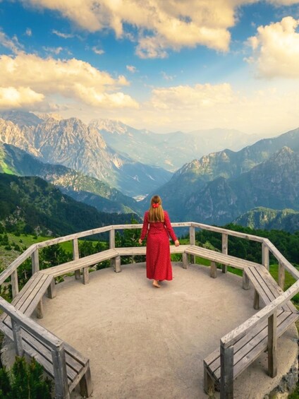 A woman in a red dress standing on a wooden platform with a view of mountains. 1