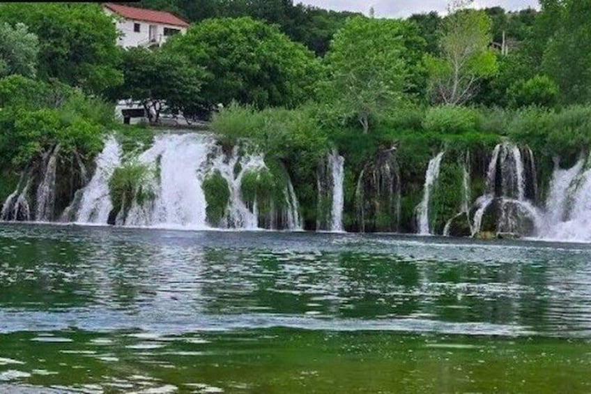 A waterfall cascading over a rocky ledge into a body of water, surrounded by lush greenery and a building in the background. 1