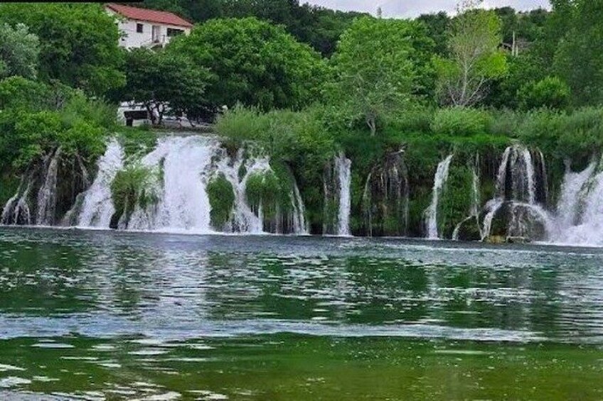 A waterfall cascading over a rocky cliff into a body of water, surrounded by lush greenery and a building in the background. 1