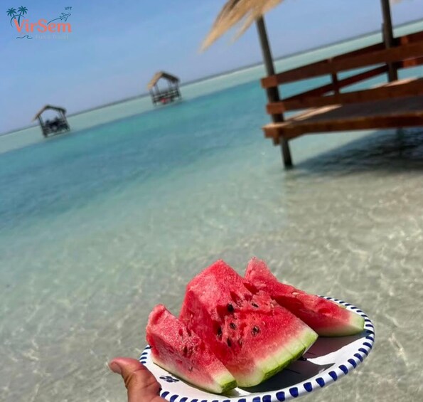 A plate of watermelon slices on a beach with clear turquoise water and wooden structures in the background. 4