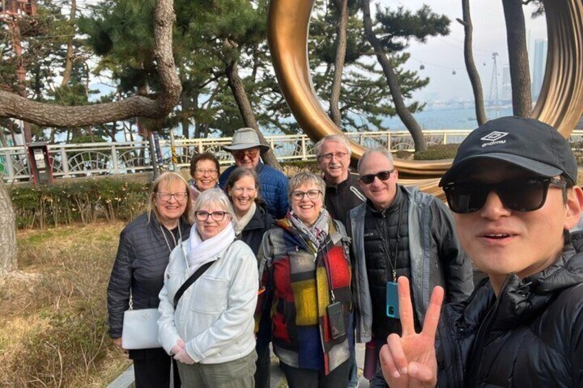 A group of people posing in front of a large, circular sculpture with a reflective surface, surrounded by trees and a body of water in the background. 5