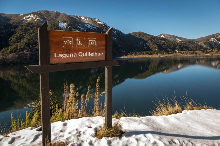 A sign for Laguna Quillihue with symbols for camping and photography, surrounded by snow, grass, and a calm lake with mountains in the background. 1