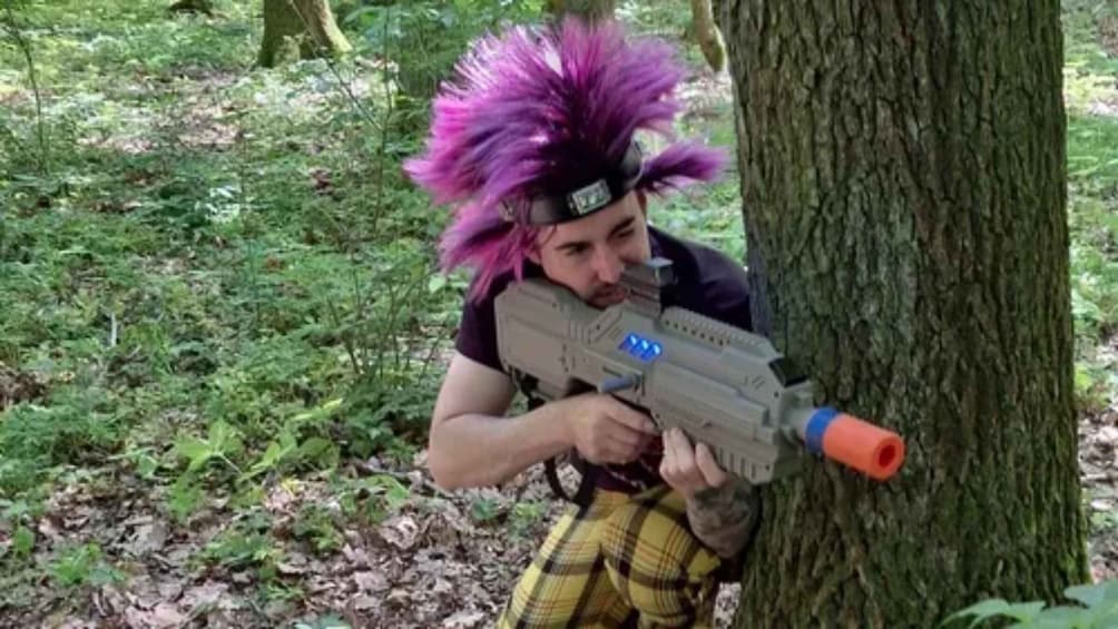 A person with purple hair and a black headband aiming a toy gun at a tree in a forest. 1