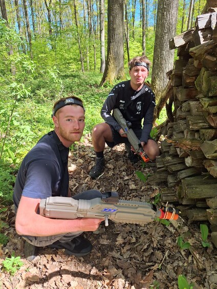 Two individuals in a forest, one holding a rifle and the other crouching beside a stack of logs. 3
