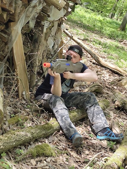 A person in camouflage clothing holding a toy gun in a wooded area with fallen logs and trees. 2