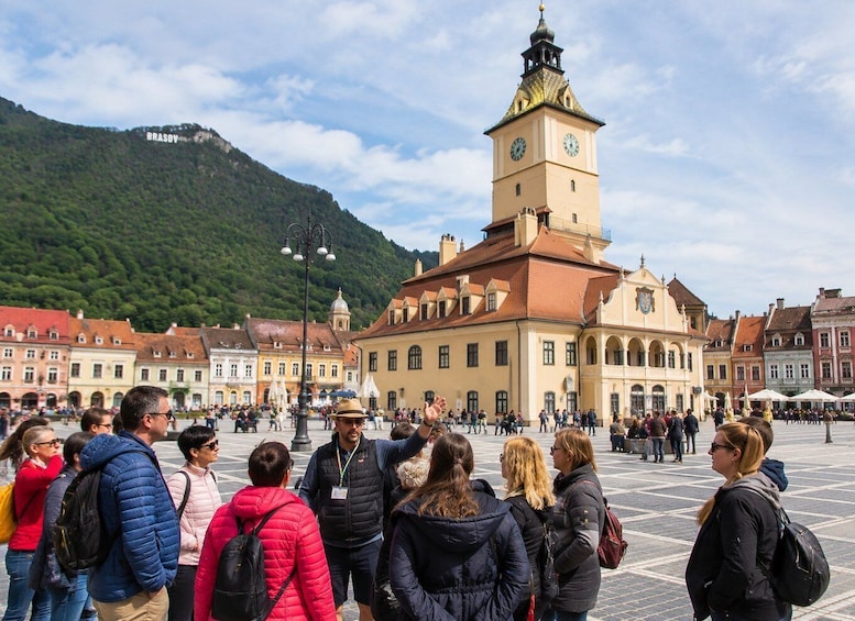 A historic square with a prominent clock tower, surrounded by traditional buildings and a mountainous backdrop. 4