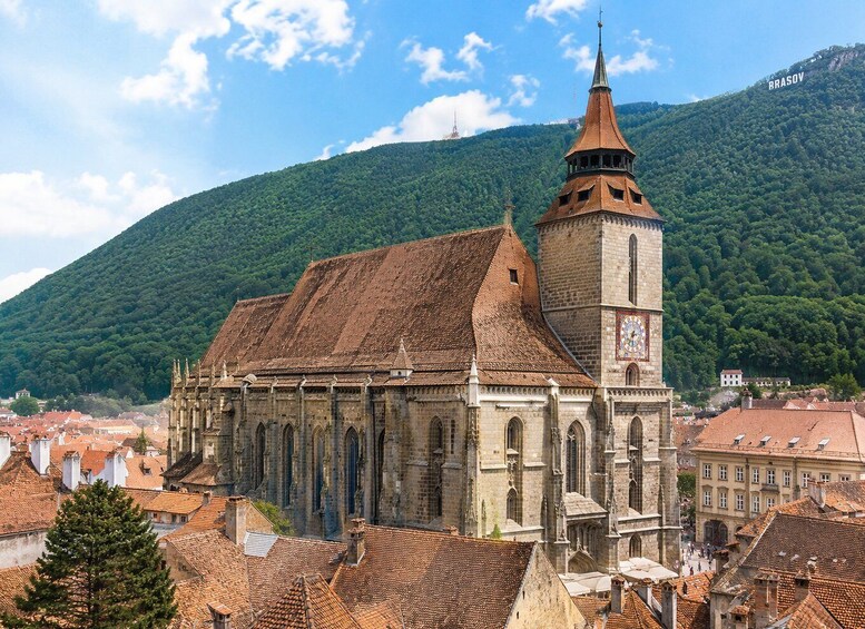 A historic church with a prominent tower and a clock, surrounded by a town with red-tiled roofs and a forested hill in the background. 2