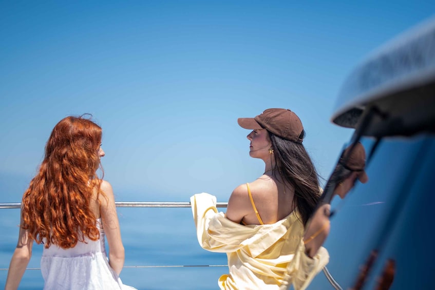 Two women on a boat, one in a white dress and the other in a yellow dress and cap, with a clear blue sky in the background. 2