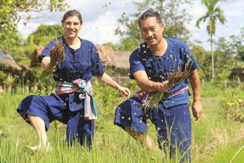 Two people in blue attire harvesting crops in a field with palm trees and thatched-roof structures in the background. 1