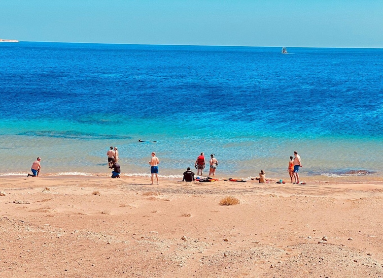 A beach with people sunbathing and swimming, a boat in the distance, and clear blue water. 3