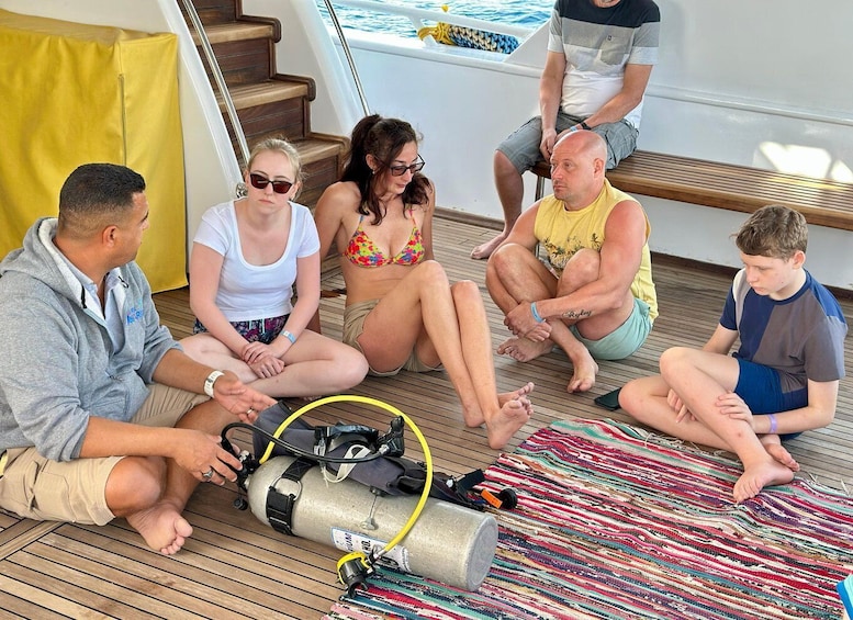 A group of people sitting on a wooden deck with scuba diving equipment on the ground. 2