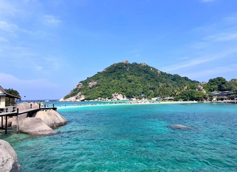 A tropical beach with clear turquoise waters, a wooden pier, and a lush green hill in the background. 3